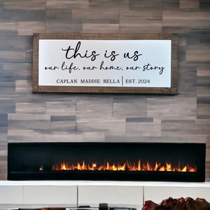 May include: A wooden sign with the text "this is us our life, our home, our story CAPLAN MADDIE BELLA | EST. 2024" hanging above a fireplace with a black surround and a roaring fire.