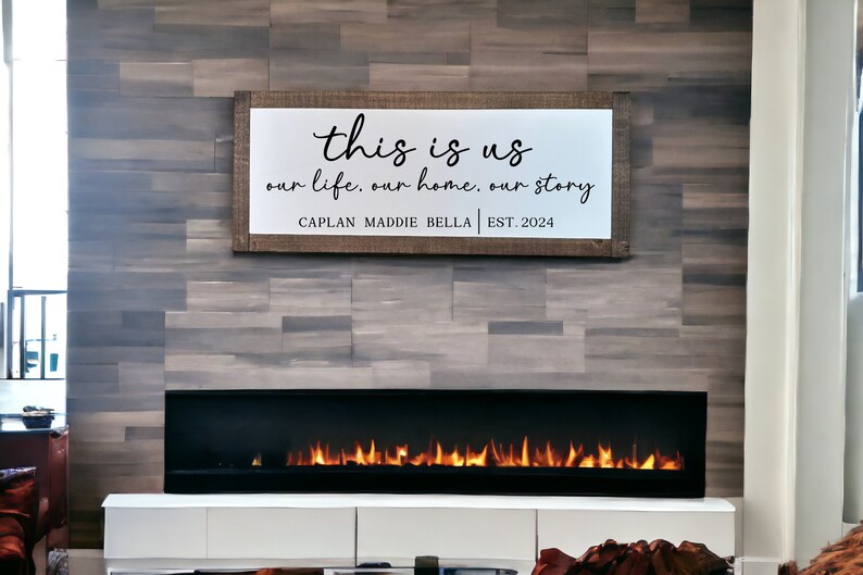 May include: A wooden sign with the text "this is us our life, our home, our story CAPLAN MADDIE BELLA | EST. 2024" hanging above a fireplace with a black surround and a roaring fire.