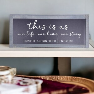 May include: A gray wooden sign with white text that reads "this is us our life. our home. our story HUNTER ALEXIS THEO | EST. 2020".