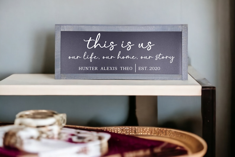 May include: A gray wooden sign with white text that reads "this is us our life. our home. our story HUNTER ALEXIS THEO | EST. 2020".
