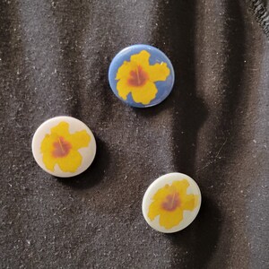 May include: Three round buttons with yellow hibiscus flowers on a black fabric background. The buttons are white with yellow flowers, white with yellow flowers, and blue with yellow flowers.