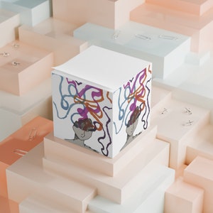 Puede incluir: Un bloc de notas blanco con un diseño abstracto colorido que presenta una cara y líneas en espiral. El bloc de notas está sentado sobre una pila de cajas blancas y beige.