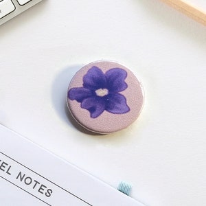 Può includere: Un bottone rotondo di colore lilla chiaro con un disegno di un singolo fiore viola.