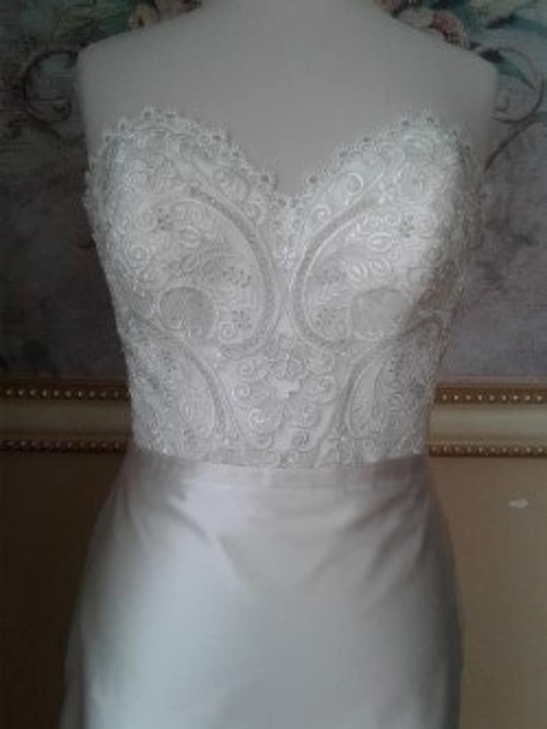 K&ouml;nnte beinhalten: Ein wei&szlig;es, tr&auml;gerloses Brautkleid mit einem figurbetonten Oberteil und einem flie&szlig;enden Rock. Das Oberteil ist mit aufwendigen Spitzenbord&uuml;ren verziert.
