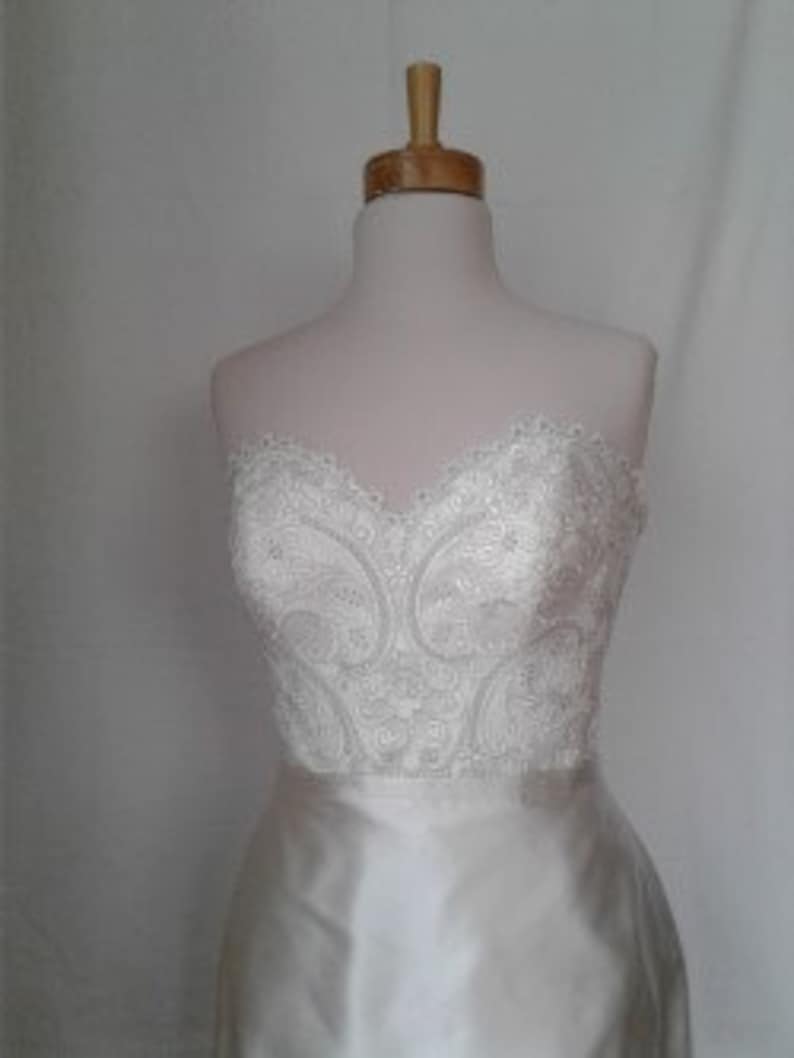 K&ouml;nnte beinhalten: Ein wei&szlig;es, tr&auml;gerloses Brautkleid mit einem Spitzenoberteil und einem Satinrock.