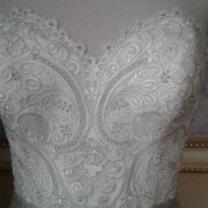 K&ouml;nnte beinhalten: Nahaufnahme eines wei&szlig;en Satin-Hochzeitskleides mit aufwendigen Spitzendetails. Die Spitze zeigt ein Paisley-inspiriertes Muster mit kleinen, klaren Perlen.