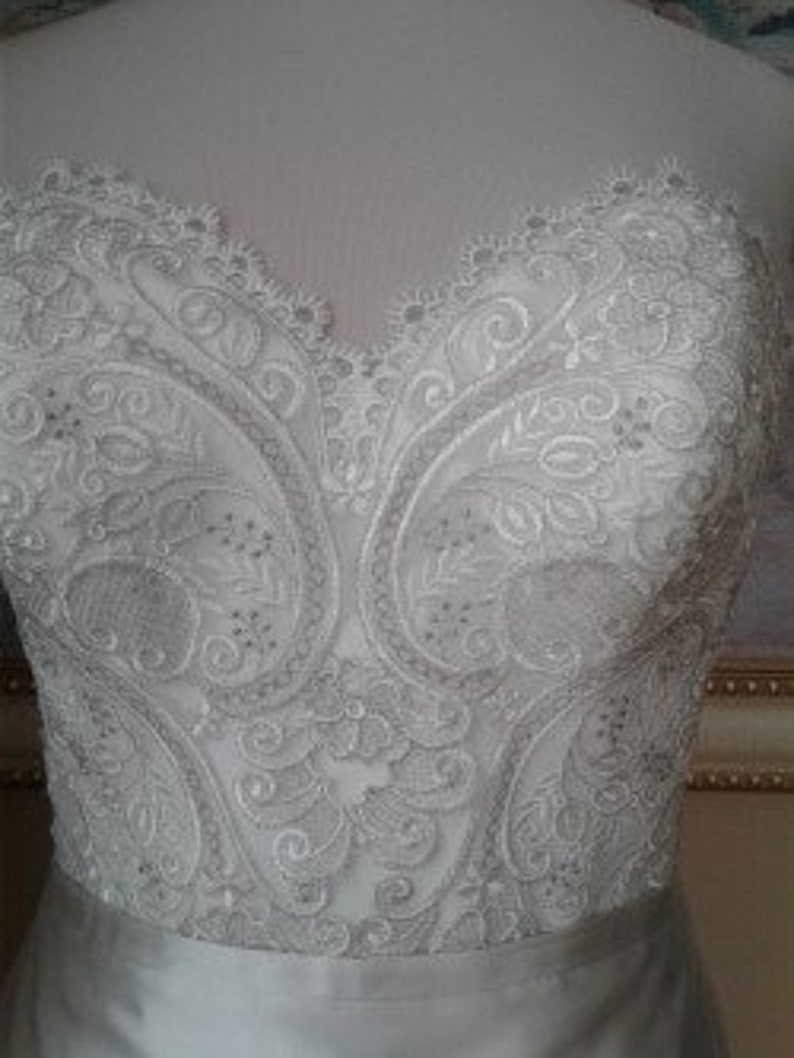 K&ouml;nnte beinhalten: Nahaufnahme eines wei&szlig;en Satin-Hochzeitskleides mit aufwendigen Spitzendetails. Die Spitze zeigt ein Paisley-inspiriertes Muster mit kleinen, klaren Perlen.