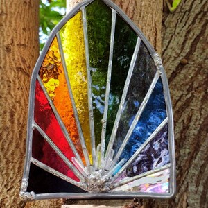 May include: A stained glass rainbow suncatcher with a silver frame. The suncatcher is made of different coloured glass pieces, including red, orange, yellow, green, blue, and purple. The suncatcher is hanging from a tree branch.