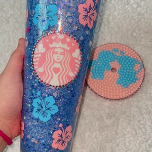 Może przedstawiać: Niebieski i r&oacute;żowy kubek z brokatem z logo Starbucks i kwiatami hibiskusa. Kubek ma pasującą pokrywkę z r&oacute;żowym i niebieskim wzorem.