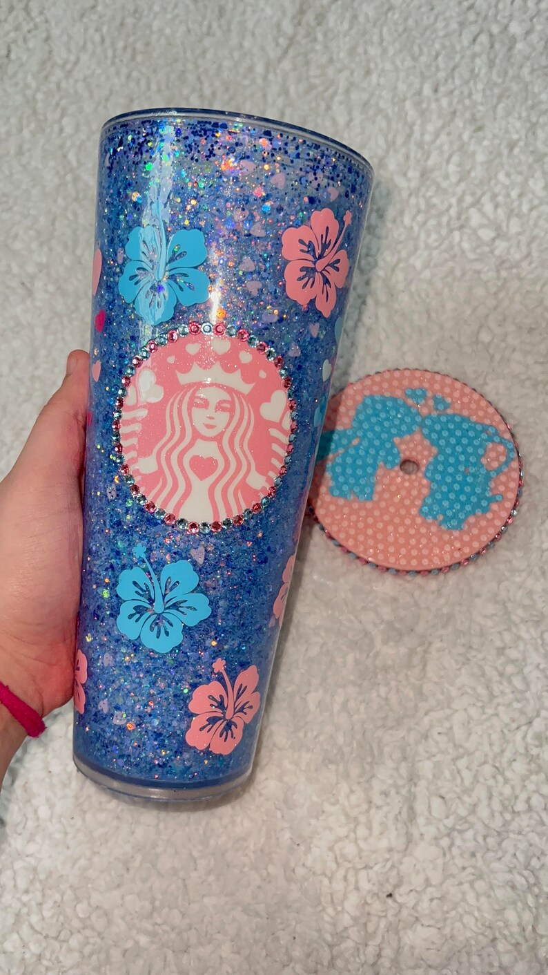 Może przedstawiać: Niebieski i r&oacute;żowy kubek z brokatem z logo Starbucks i kwiatami hibiskusa. Kubek ma pasującą pokrywkę z r&oacute;żowym i niebieskim wzorem.