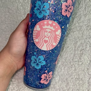 Może przedstawiać: Przezroczysty, wielokrotnego użytku kubek z niebiesko-r&oacute;żową podstawą z brokatem. Kubek zdobi r&oacute;żowe logo Starbucks z sercami i koroną oraz niebieskie i r&oacute;żowe kwiaty hibiskusa.