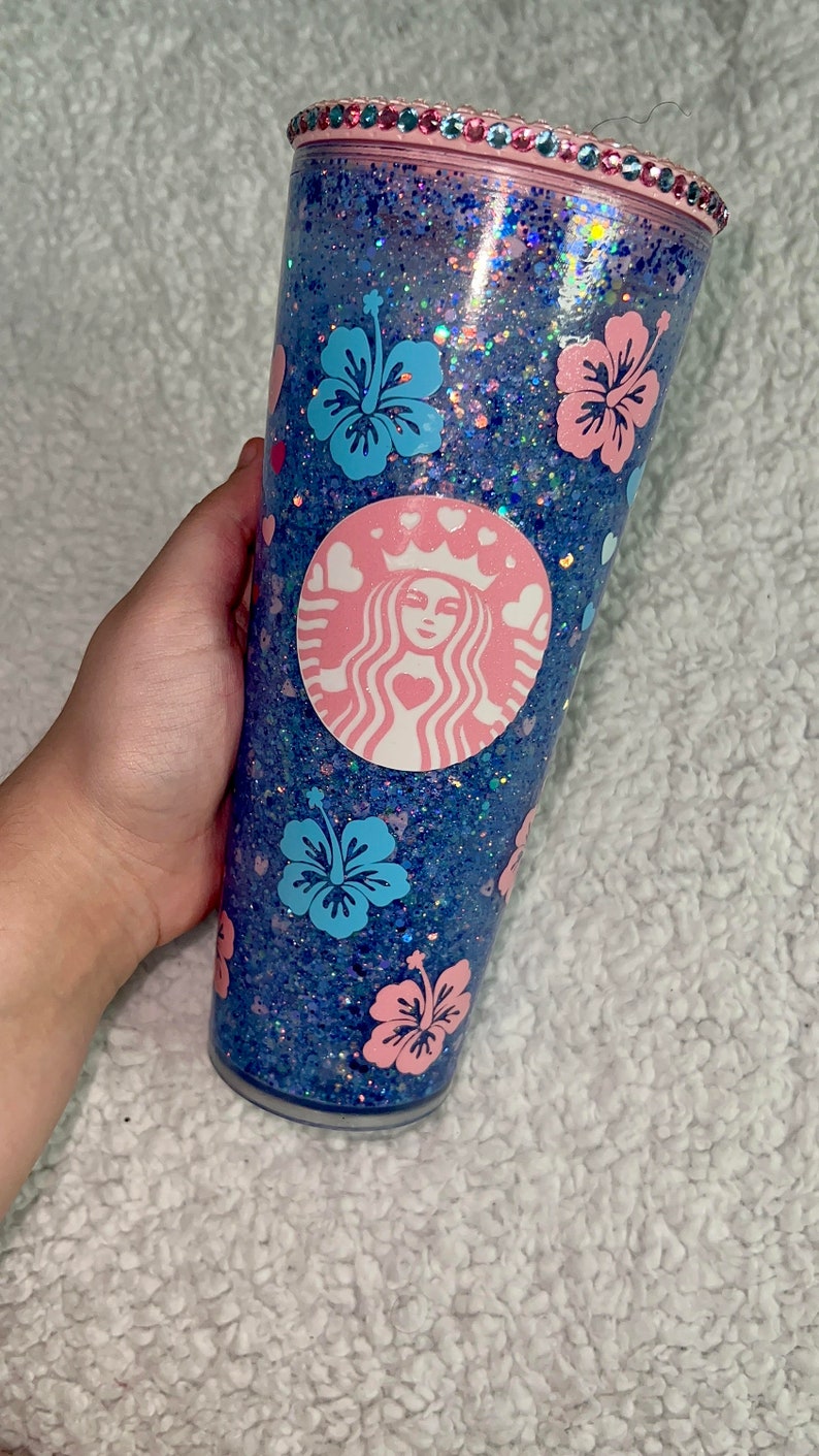 Może przedstawiać: Przezroczysty, wielokrotnego użytku kubek z niebiesko-r&oacute;żową podstawą z brokatem. Kubek zdobi r&oacute;żowe logo Starbucks z sercami i koroną oraz niebieskie i r&oacute;żowe kwiaty hibiskusa.