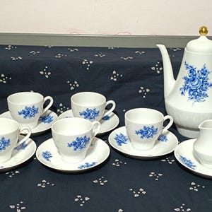 Pode incluir: Um conjunto de chá em porcelana branca com um padrão floral azul. O conjunto inclui um bule alto com um remate dourado na tampa, uma pequena leiteira e quatro chávenas com pires a condizer, decorados com motivos de rosas e folhas azuis.