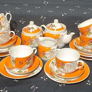 Könnte beinhalten: Ein Teeservice bestehend aus einer Teekanne, einer Zuckerdose mit Deckel, einem Milchkännchen sowie vier Tassen mit passenden Untertassen und Tellern. Jedes Teil ist weiß mit leuchtend orangefarbenen Flächen und einem botanischen Muster.
