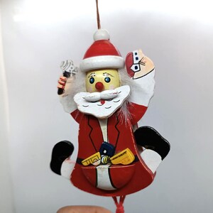 May include: A hanging wooden Santa Claus ornament painted in red, white, and black. The figure features a red hat, a white beard, and a red coat with a pocket containing small yellow and blue toy shapes. One hand holds a small white object.