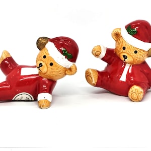 Puede incluir: Dos figuras de osos de peluche de cerámica con gorros de Papá Noel rojos y trajes rojos. Un oso está tumbado de lado con una pierna levantada, mientras que el otro está sentado con un brazo y una pierna extendidos. Ambos tienen pelaje beige.