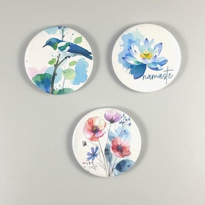Könnte beinhalten: Drei runde Untersetzer aus Keramik mit Aquarell-Blumen- und Vogelmotiven. Einer zeigt einen blauen Vogel auf einem Ast, ein anderer eine blaue Lotusblume mit dem Wort "namaste" und der dritte bunte Blumen. Jeder Untersetzer hat einen weißen Hintergrund.
