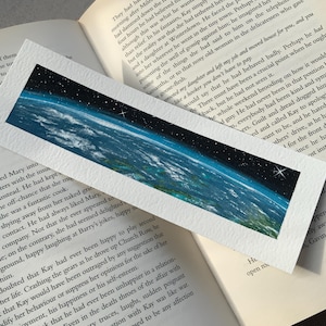May include: A rectangular bookmark with a painted image of Earth from space, showing the curvature of the planet, blue oceans, and a starry sky. The bookmark is resting on an open book.