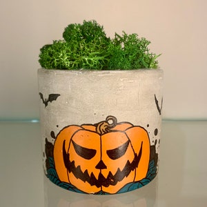 May include: A cylindrical gray concrete planter featuring a painted orange jack-o'-lantern with a jagged black face, surrounded by black bat silhouettes and blue leaf accents, filled with a cluster of vibrant green preserved reindeer moss.