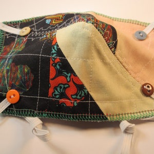 May include: A black, green, red, and orange patterned fabric face mask with a beige fabric panel and a peach colored fabric panel. The mask has white elastic straps and four buttons.
