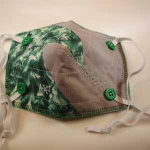 May include: A green and gray fabric face mask with white elastic straps and green buttons. The mask is quilted and has a snowflake pattern.