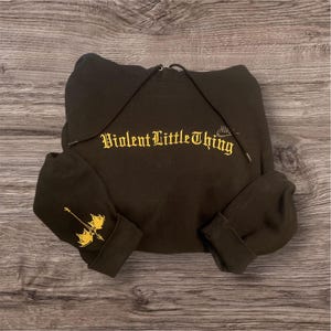 Könnte beinhalten: Schwarzer Kapuzenpullover mit dem goldenen Schriftzug "Violent Little Thing" auf der Brust. Am linken Ärmelaufschlag befindet sich eine goldene Grafik einer Waffe. Der Hoodie hat eine Kapuze mit Kordelzug.