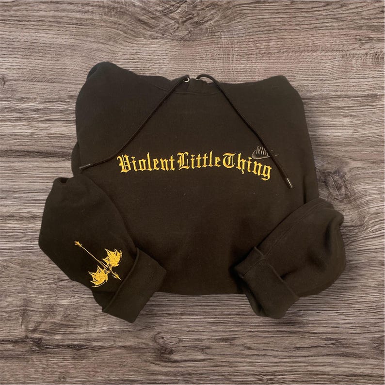 May include: Black hoodie with the text "Violent Little Thing" in gold lettering across the chest. The left sleeve cuff features a gold graphic of a weapon. The hoodie has a drawstring hood.