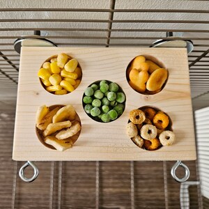 May include: A wooden bird feeder with five compartments filled with different types of bird food. The feeder is attached to a cage with metal hooks.