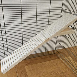 May include: A light-colored wooden ramp with grooved steps, designed for a small animal cage. The ramp is angled and attached to the cage's wire mesh. The cage is set on a wooden floor.