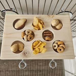 May include: A wooden bird feeder with suction cups for attaching to a cage. The feeder has nine compartments filled with different types of birdseed and nuts.