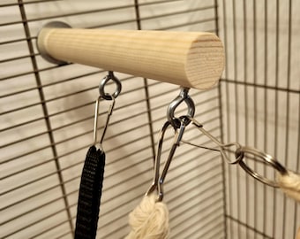 Pode incluir: Um poleiro de madeira para pássaros com ferragens de metal, incluindo uma alça preta e um brinquedo de corda, preso a uma gaiola. O poleiro é cilíndrico e feito de madeira clara. As barras da gaiola são visíveis no fundo.