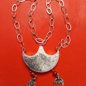Peut inclure: Collier argenté avec une chaîne à maillons ovales et un pendentif en forme de croissant. Le pendentif présente des motifs floraux gravés et deux breloques pendantes avec des perles rouges. Sur fond rouge.