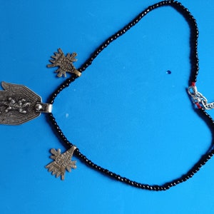 Puede incluir: Un collar de cuentas negras con tres colgantes de plata. Los colgantes son una mano con un escorpión, un árbol estilizado y otro árbol estilizado.