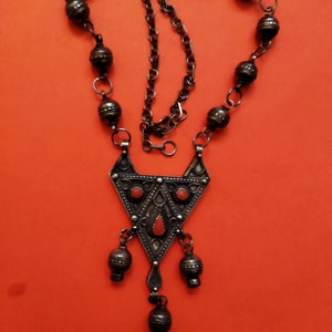 May include: A silver necklace with a triangular pendant featuring red stones and intricate details. The pendant hangs from a chain with silver beads.