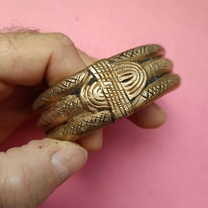 May include: A gold-colored metal bracelet with intricate woven designs. The bracelet is made of three bands with a raised, textured design.