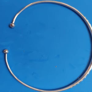 Könnte beinhalten: Eine silberfarbene, offene Halskette bestehend aus einem dünnen, kreisförmigen Metallband, das an beiden Enden in einer kleinen, glatten, kugelförmigen Perle endet. Das Design ist flexibel und bildet einen weiten C-Bogen mit polierter Oberfläche.