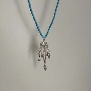 Può includere: Una collana con un ciondolo a forma di mano di Hamsa in argento e una catena di perline blu. La mano di Hamsa è un simbolo popolare di protezione e buona fortuna.