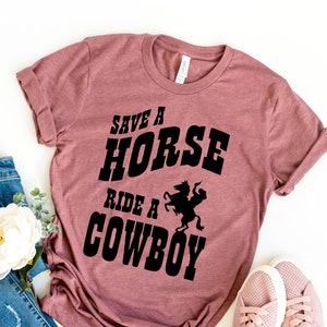 May include: A mauve t-shirt with the text "Save a Horse Ride a Cowboy" in black lettering. The shirt features a silhouette of a cowboy riding a horse.