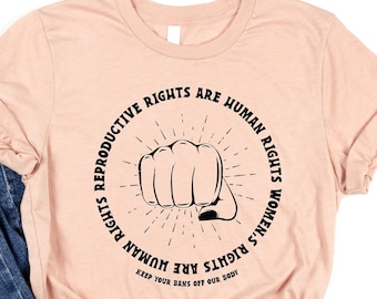 Könnte beinhalten: Ein pfirsichfarbenes T-Shirt mit einer schwarzen Grafik einer Faust und dem Text "Reproductive Rights Are Human Rights Women's Rights Are Human Rights Keep Your Hands Off Our Body".