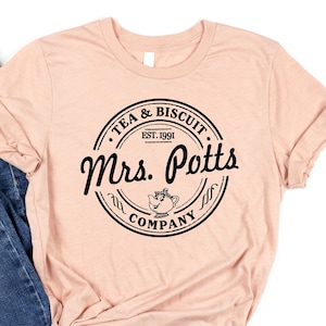 May include: A peach-colored t-shirt with a black circle design that says "Tea & Biscuit Company" with a teapot and the text "Mrs. Potts" in a script font.
