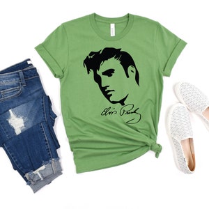 May include: A green t-shirt with a black silhouette of Elvis Presley's face and his signature "Elvis Presley". The t-shirt is laid flat on a white background with a pair of white striped canvas shoes and a pair of blue jeans.