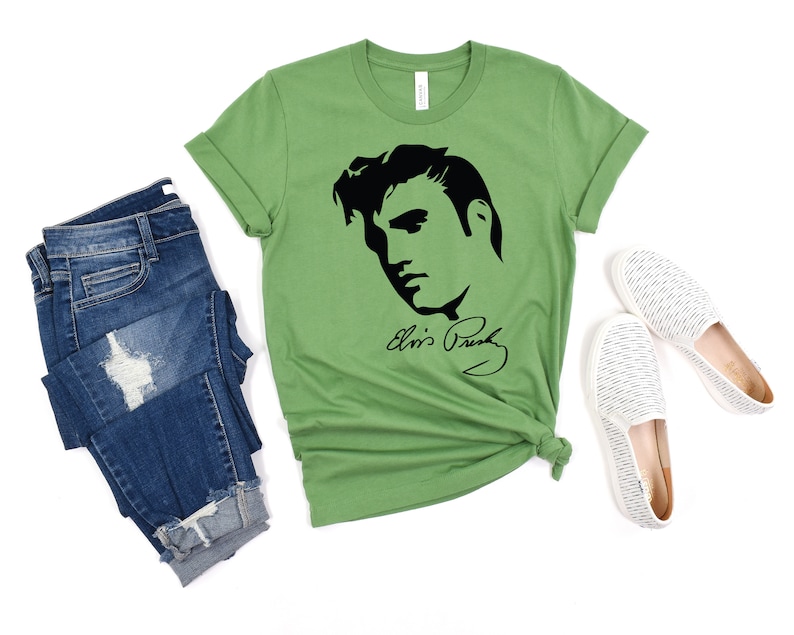 May include: A green t-shirt with a black silhouette of Elvis Presley's face and his signature "Elvis Presley". The t-shirt is laid flat on a white background with a pair of white striped canvas shoes and a pair of blue jeans.