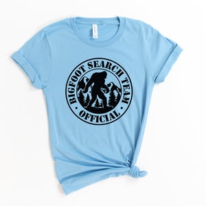 May include: A light blue t-shirt with a black circle graphic that says "Bigfoot Search Team Official" with a silhouette of Bigfoot walking in the mountains.