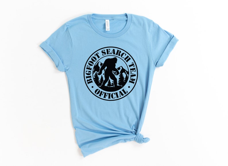 May include: A light blue t-shirt with a black circle graphic that says "Bigfoot Search Team Official" with a silhouette of Bigfoot walking in the mountains.