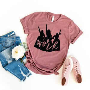 Peut inclure: Un t-shirt mauve avec une silhouette noire de trois femmes et le texte "work" en noir.