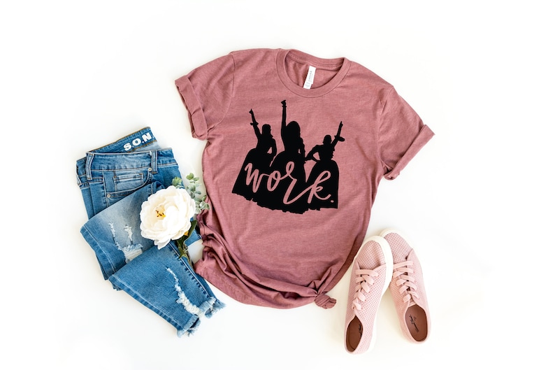 Peut inclure: Un t-shirt mauve avec une silhouette noire de trois femmes et le texte "work" en noir.