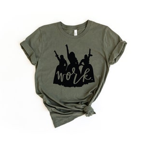 Peut inclure: Un t-shirt vert avec une silhouette noire de trois femmes en robes et le mot "work" &eacute;crit en cursive noire.