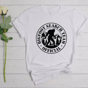 May include: White t-shirt with a black circle graphic that says "Bigfoot Search Team Official" with a silhouette of Bigfoot walking in the mountains.