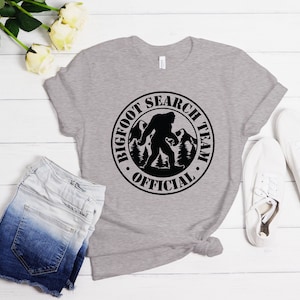 May include: A gray t-shirt with a black circle graphic that says "Bigfoot Search Team Official" with a silhouette of Bigfoot walking in the woods.