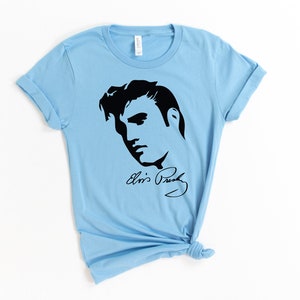 May include: Light blue t-shirt with a black silhouette of Elvis Presley's face and his signature "Elvis Presley".